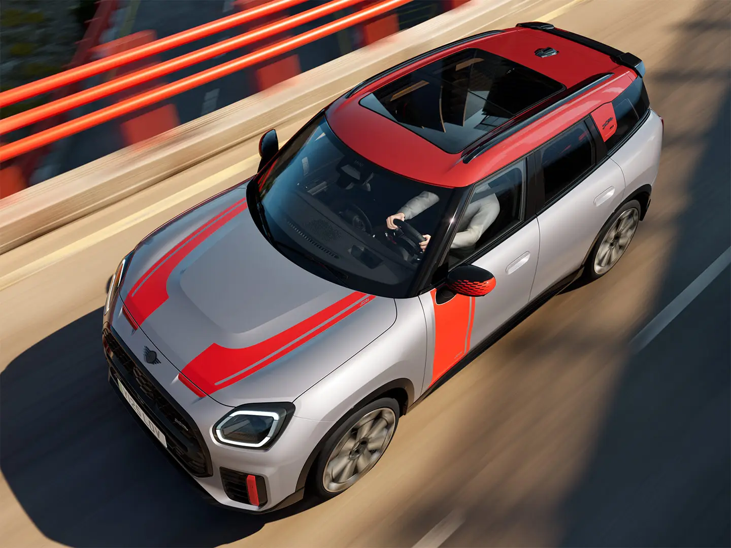MINI Countryman John Cooper Works in Melting Silver with mirror cap, side and bonnet decals, roof, and winglet in Chili Red, driving along city street – front-side view.