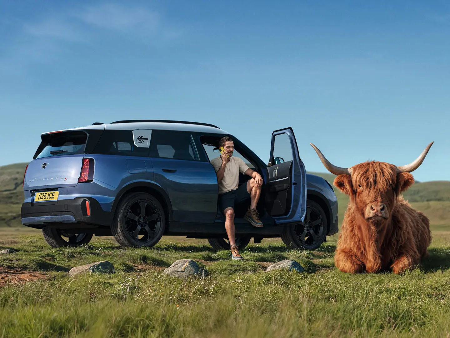 Man outside his MINI Countryman S talking to MINI Roadside Assistance via the MINI App with a  Highland Bull lying nearby.